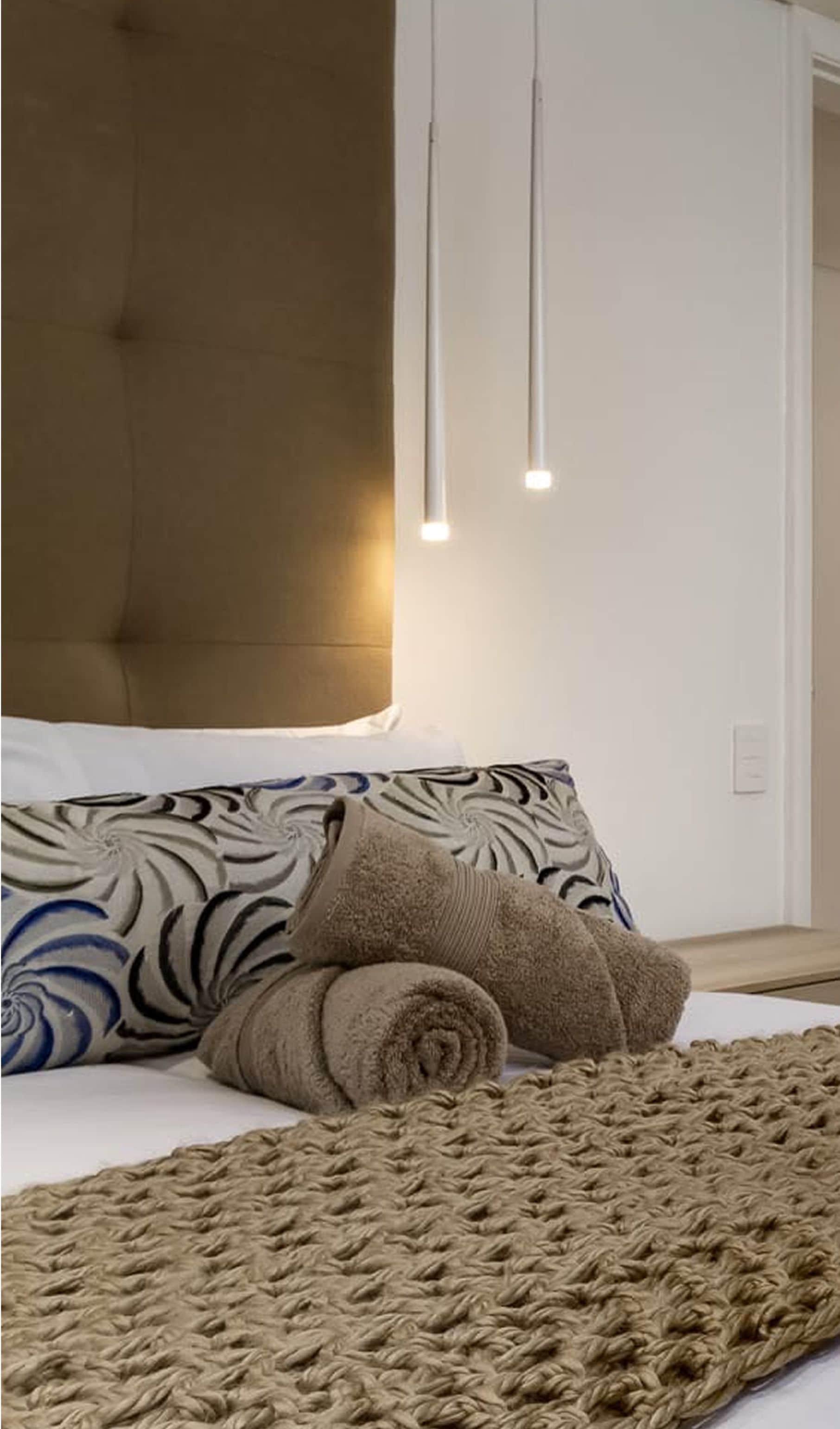 Guest bed with rolled towels, a swirl-patterned cushion and slim pendant lights beside an upholstered headboard