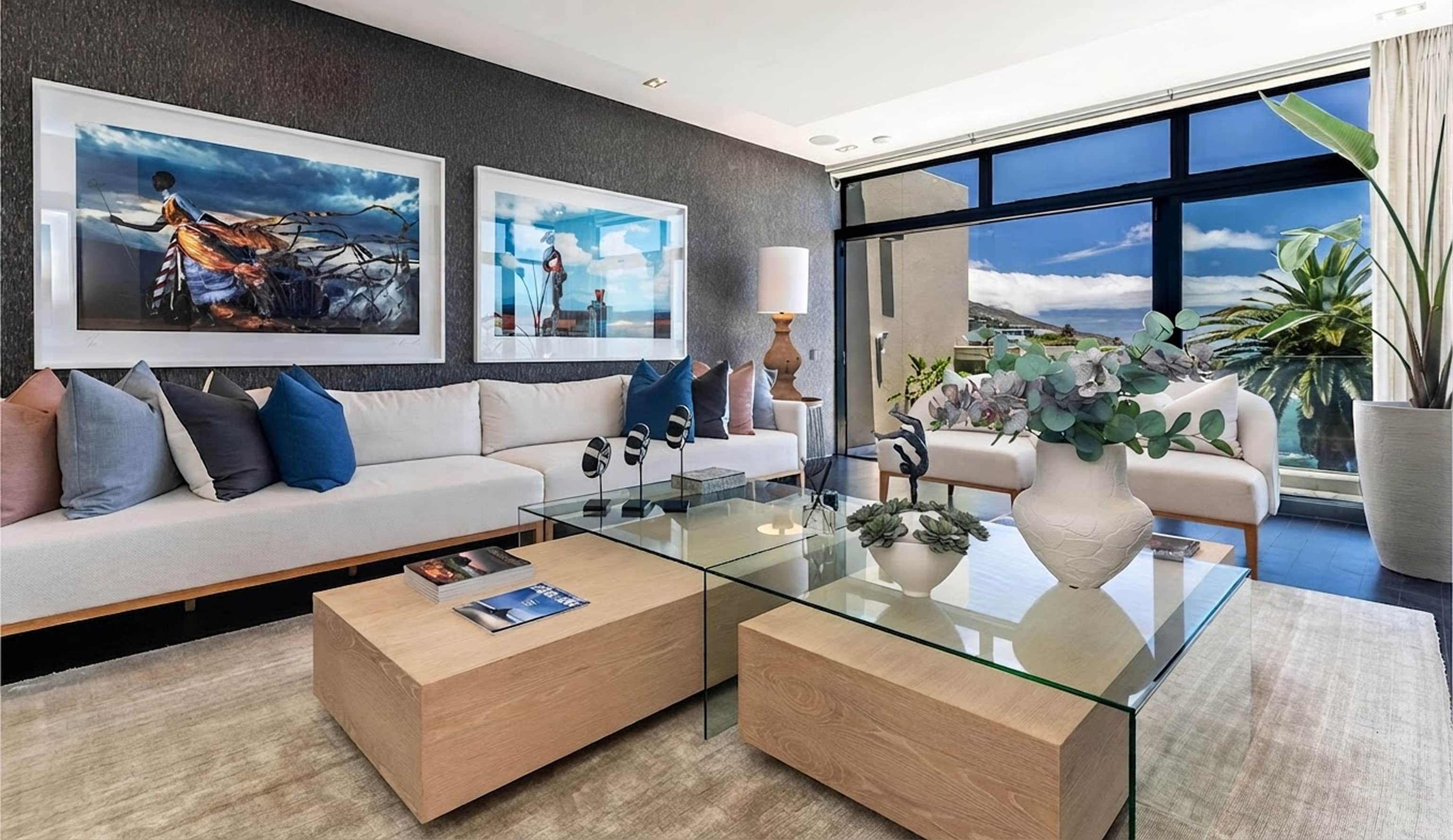 Lounge with a white sofa, blue cushions, framed photographs and glass-and-oak coffee tables beside tall windows