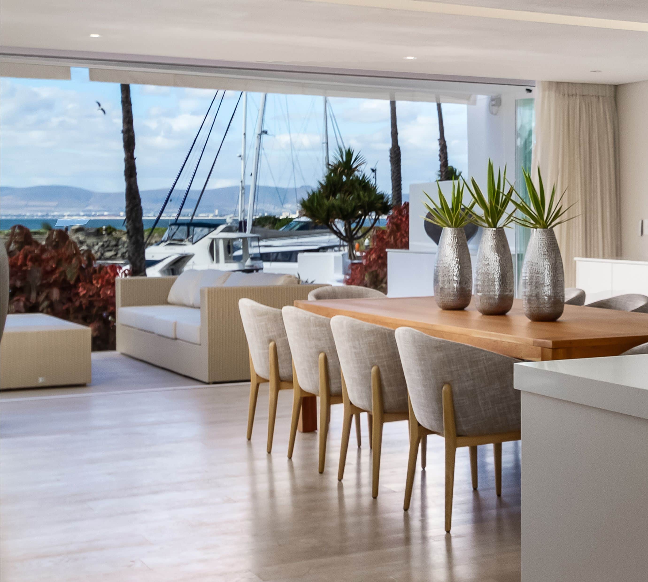 Dining table with upholstered chairs and silver vases, opening onto a terrace overlooking a marina