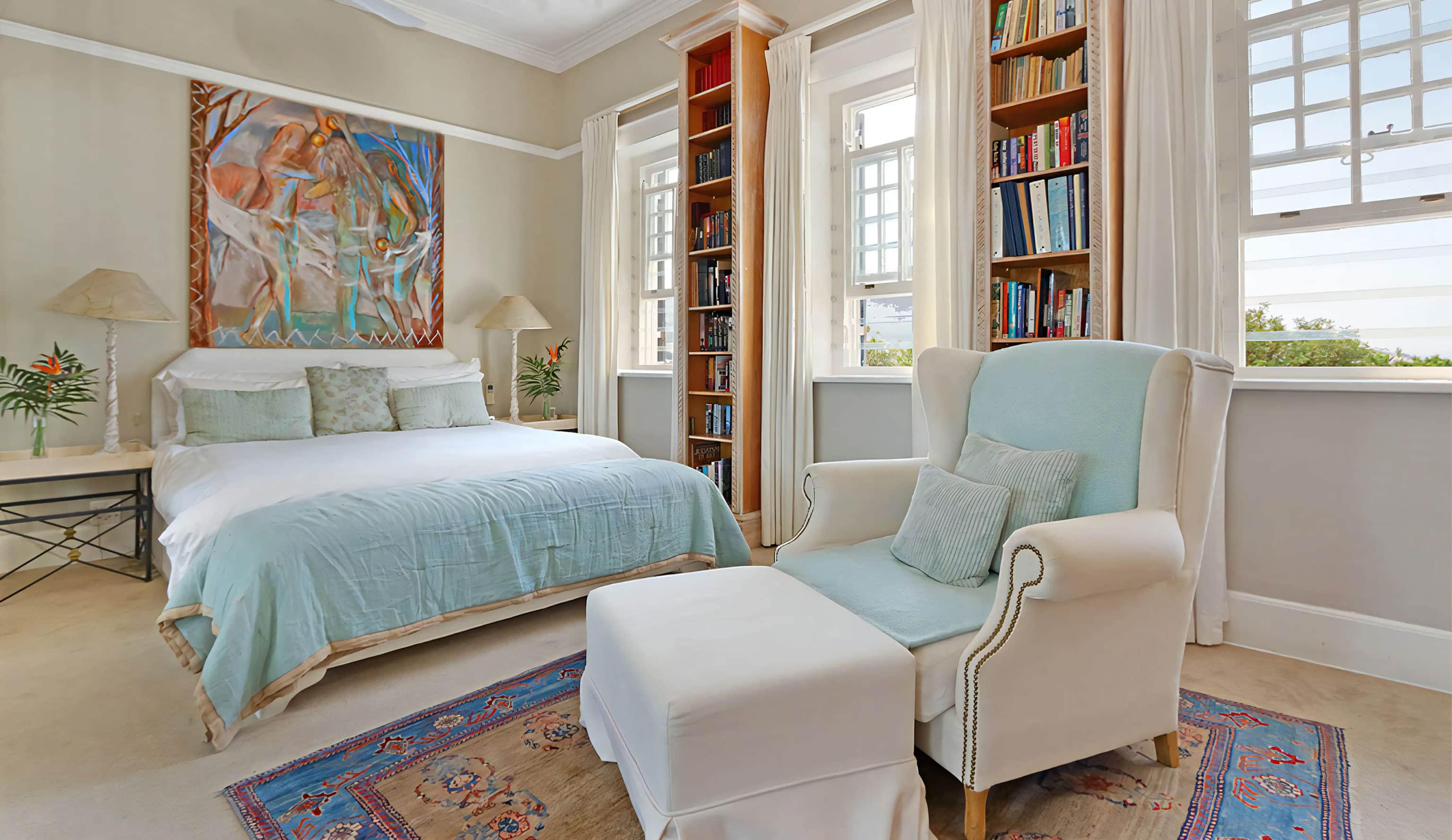 Bedroom with a pale blue bedspread, a wingback armchair and footstool, and bookshelves between sash windows