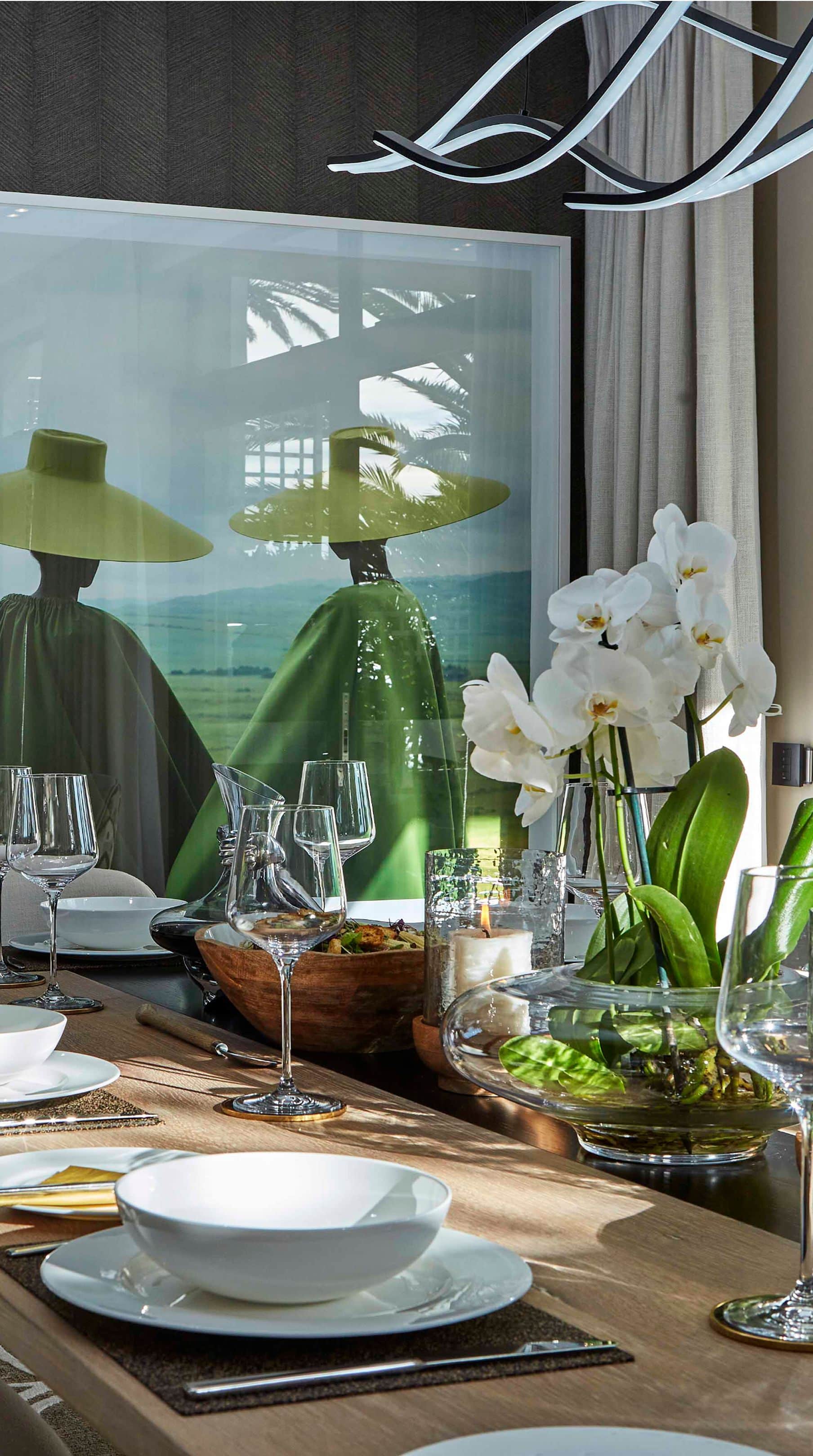 Dining table set with white plates, glassware and orchids beneath a photograph of figures in green hats
