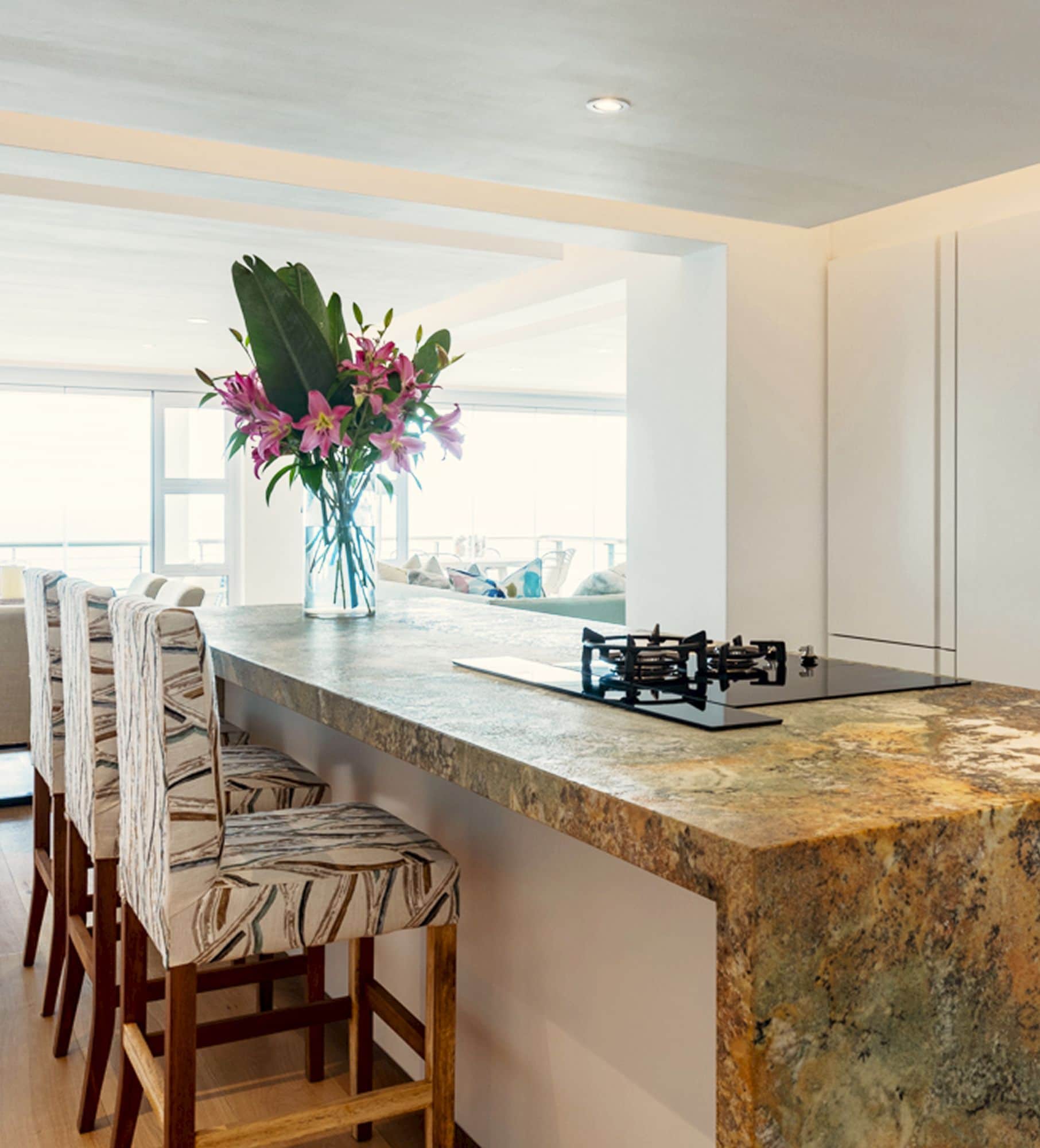 Kitchen island with a granite counter, gas hob and patterned bar stools, open to a sea-facing lounge