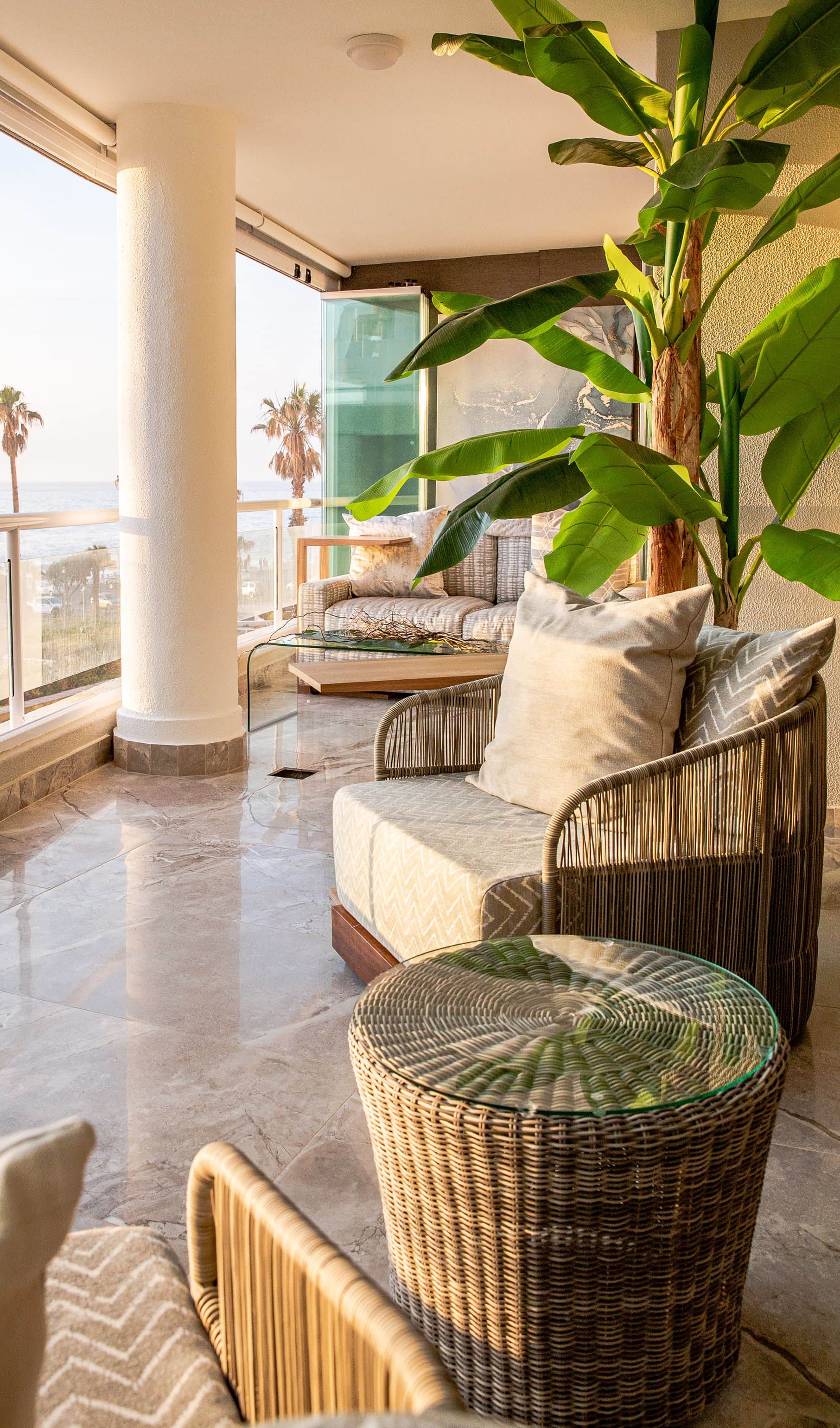 Covered terrace with a banana plant, a woven armchair and a glass-topped side table overlooking the sea