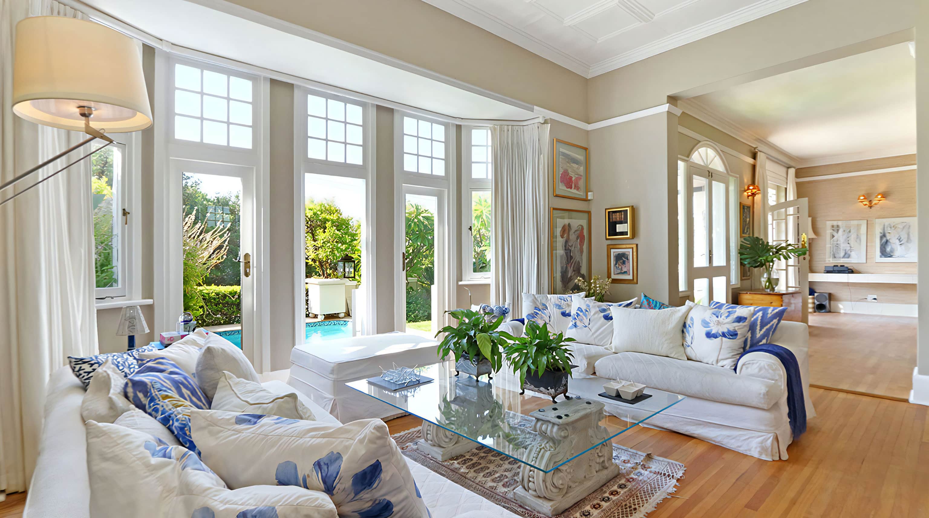 Living room with tall bay windows overlooking the pool, white sofas and a glass coffee table